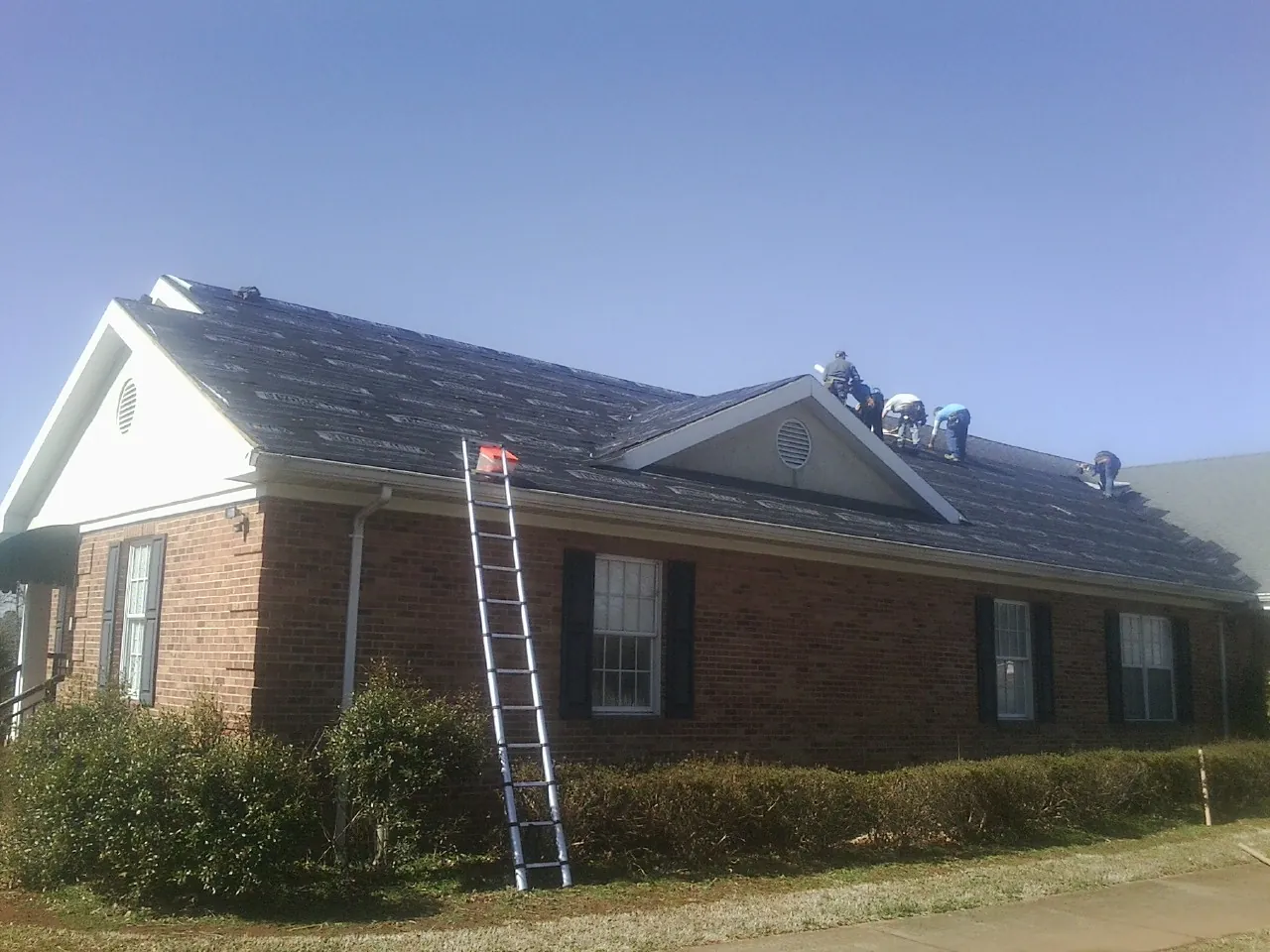Crew tearing shingles off a brick ranch roof with a ladder set — Metal Roof Repair work by our crew in Emigrant