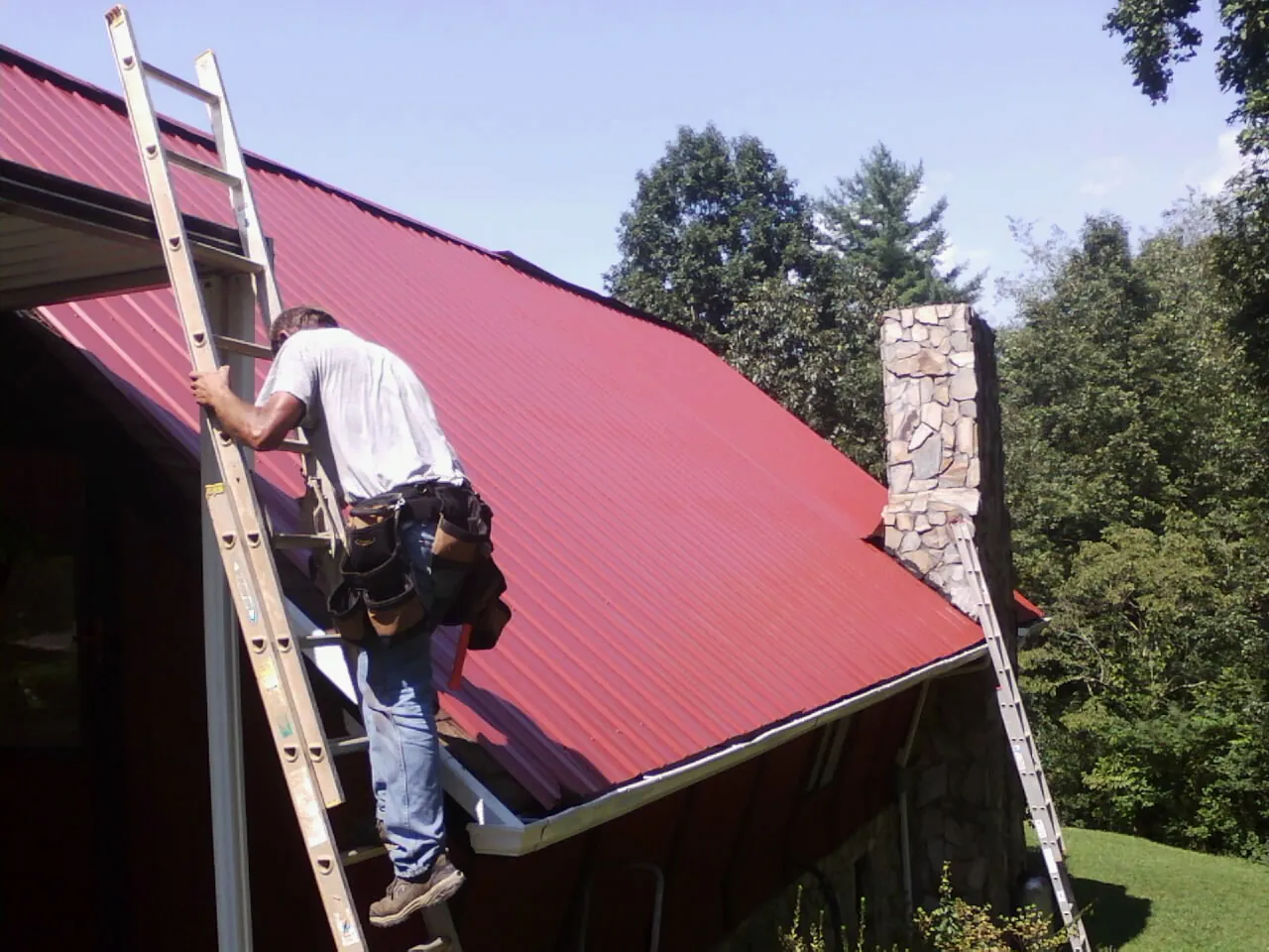 Roofer on a ladder beside a red metal roof and stone chimney — Soffit & Fascia Repair by Roofing Brothers in Emigrant